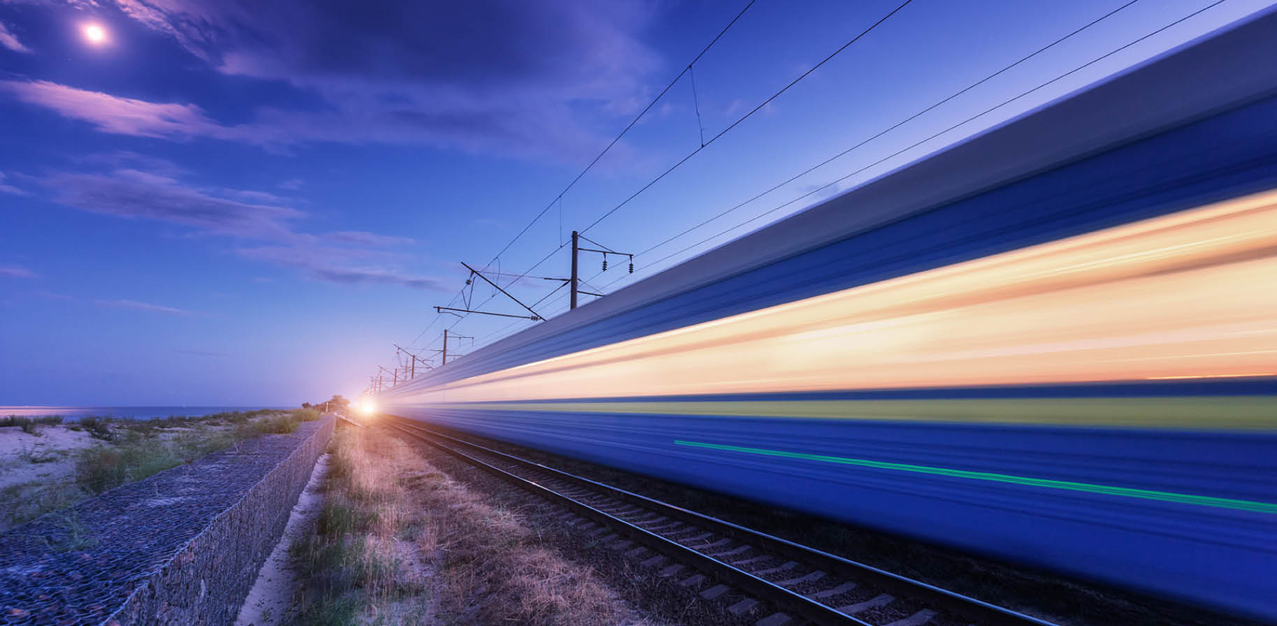 High speed passenger train in motion on the railroad at summer night. Moving blurred modern commuter train at dusk. Railway station and purple sky with moon and clouds. Industrial landscape. Transport